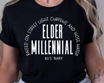 May include: Black t-shirt with white text that reads "Raised on street lights, curfews and hose water. Elder Millennial. 80's Baby."