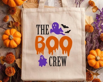 May include: A beige canvas tote bag featuring the text "THE BOO CREW" in black, orange, and purple, with ghost and bat graphics. The bag is set amongst orange pumpkins, dried leaves, and purple flowers, evoking a Halloween atmosphere.
