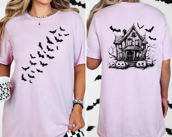 May include: A light pink t-shirt featuring a black bat design on the front and a haunted house graphic on the back. The shirt has short sleeves and a crew neck. The design is perfect for Halloween.