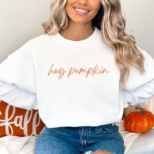 Puede incluir: Una sudadera blanca con la frase "hey pumpkin" en escritura color cobre. La sudadera se combina con vaqueros azules. Una calabaza naranja y un cojín con temática otoñal están en el fondo.