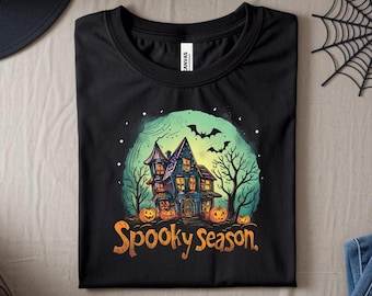May include: Black t-shirt featuring a colorful Halloween design. The graphic includes a haunted house, pumpkins, bats, and the text "Spooky season." The design is set against a green and blue background.
