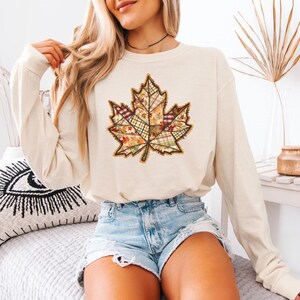 May include: Cream-colored long-sleeve shirt featuring a large, detailed maple leaf design. The leaf is composed of various fabric patterns in fall colors like orange, green, and brown. The shirt is paired with denim shorts, suggesting a casual, seasonal style.