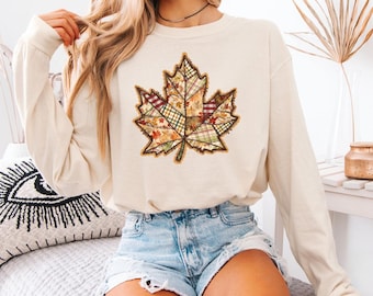 Puede incluir: Camiseta de manga larga color crema con un gran diseño de hoja de arce. La hoja está compuesta por varios patrones de tela en colores otoñales como naranja, verde y marrón. La camiseta se combina con pantalones cortos de mezclilla, lo que sugiere un estilo casual y de temporada.