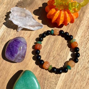 May include: An assortment of decorative items including a black beaded bracelet with orange and green accents, a purple amethyst stone, a green stone, a clear crystal, and an orange glass pumpkin.