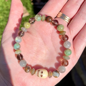 May include: A beaded bracelet featuring a skull charm and a variety of gemstone beads in shades of green, brown, and clear. The bracelet is held in a hand, showcasing the details of the beads and the skull.