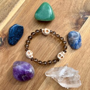 May include: A beaded bracelet with skull charms is surrounded by various gemstones. The bracelet features brown beads and white skull charms. Gemstones include a green triangle, a blue stone, a purple stone, and a clear quartz cluster.