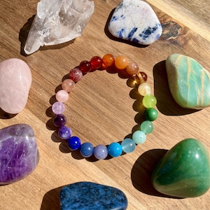 May include: A rainbow chakra bracelet made with natural gemstones. The bracelet features beads in the colors of the rainbow, including red, orange, yellow, green, blue, indigo, and violet. The bracelet is surrounded by various crystals, including clear quartz, rose quartz, amethyst, and green aventurine.
