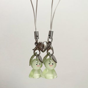 May include: A white phone charm with two green bunny figures attached to a silver heart charm. The charm is attached to a white string.