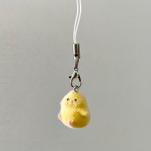 May include: A yellow felt chick keychain with black eyes and pink feet. The chick is attached to a silver clip and a white string.