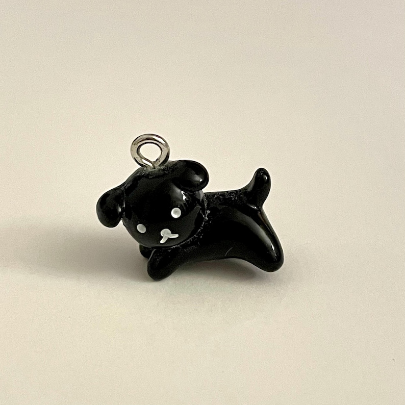 May include: Black charm of a cartoon dog with white eyes and a nose. The charm has a silver loop for attaching to a necklace or bracelet.