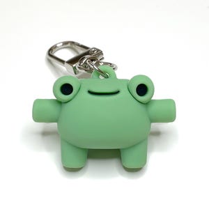May include: A light green silicone frog keychain featuring a smooth, matte finish, two prominent eyes with dark centers, a simple curved mouth, and a silver-toned metal lobster clasp attached to a small loop at the top of the frog's head.