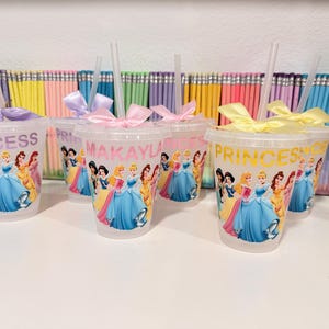 May include: A set of clear plastic tumblers with lids and straws, each decorated with colorful Disney princess graphics. Some cups feature the text "PRINCESS" or a name like "MAKAYLA" in colored lettering. Each lid is adorned with a satin ribbon bow.