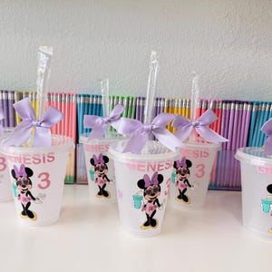 May include: A set of clear plastic cups with lids and straws, each decorated with a graphic of a cartoon mouse wearing heart-shaped glasses, the name Genesis, and the number 3. Each cup is adorned with a light purple satin ribbon bow.