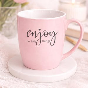 May include: A pink ceramic mug with a white interior and a curved handle. The mug features the words "enjoy the little things" in black script. It sits on a white coaster, with a blurred background of flowers and a candle.