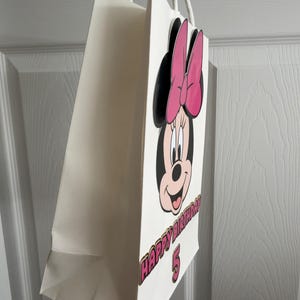 May include: A white paper gift bag featuring a cartoon image of Minnie Mouse with a pink bow. The bag has the text "HAPPY BIRTHDAY 5" in pink and gold lettering. White rope handles are attached to the top of the bag.