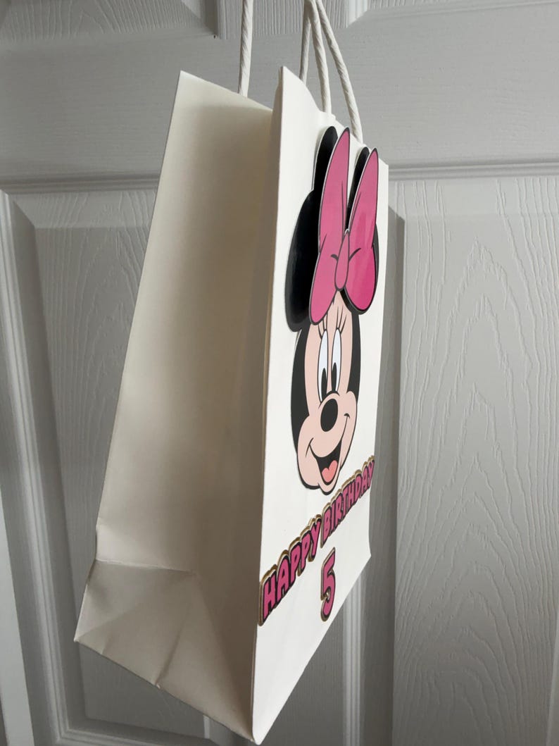 May include: A white paper gift bag featuring a cartoon image of Minnie Mouse with a pink bow. The bag has the text "HAPPY BIRTHDAY 5" in pink and gold lettering. White rope handles are attached to the top of the bag.