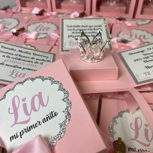 May include: Pink favor boxes with clear lids, each containing a small silver crown and a card with the name "Lia" and the text "mi primer a&ntilde;ito".