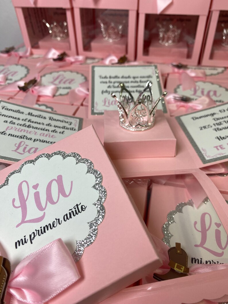 May include: Pink favor boxes with clear lids, each containing a small silver crown and a card with the name "Lia" and the text "mi primer a&ntilde;ito".