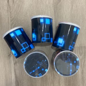 May include: Four cylindrical containers with black exteriors and blue geometric designs. The containers have white rims and clear lids, also featuring blue geometric patterns. The containers are arranged on a wooden surface.