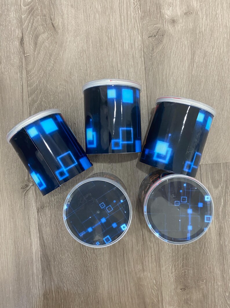 May include: Four cylindrical containers with black exteriors and blue geometric designs. The containers have white rims and clear lids, also featuring blue geometric patterns. The containers are arranged on a wooden surface.