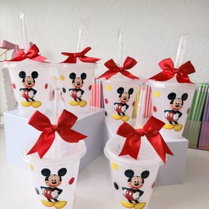 May include: Six clear plastic tumblers with lids and straws, each featuring a graphic of Mickey Mouse. The cups are decorated with red and yellow polka dots and topped with a large, bright red satin ribbon bow tied around the straw.