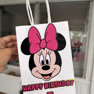 May include: White gift bag featuring Minnie Mouse with a pink bow and black ears. The bag has the text "HAPPY BIRTHDAY" in gold and pink, with the number "5" below. The bag has a white handle.