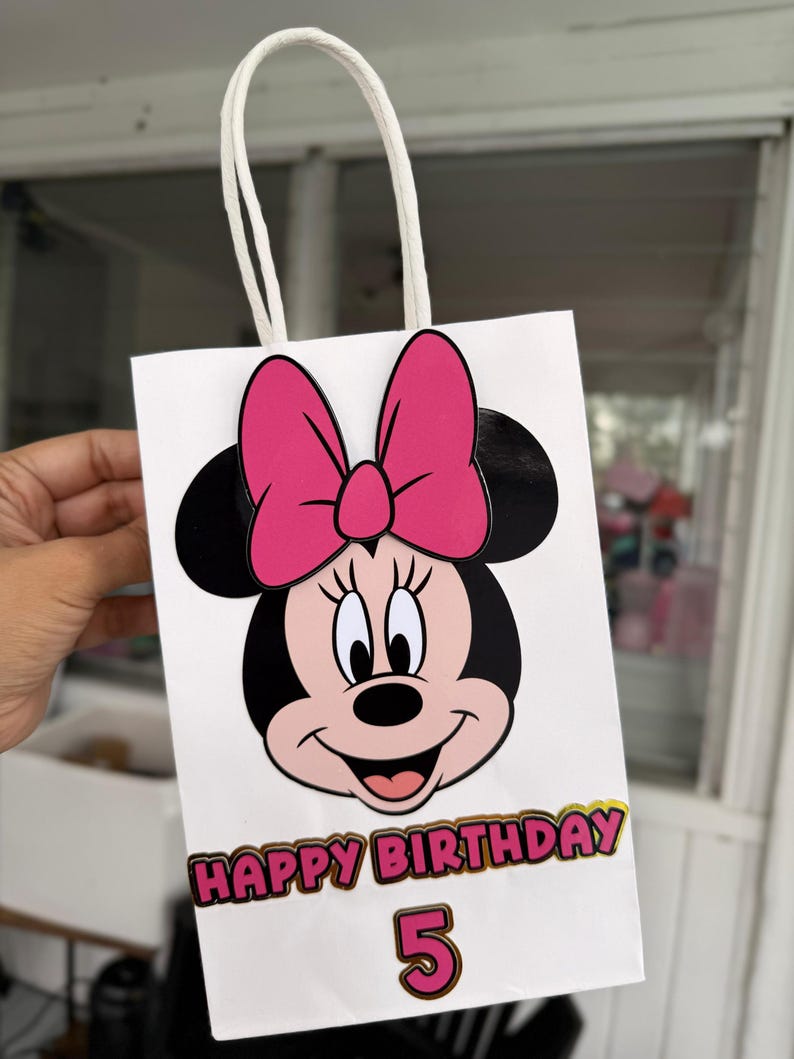 May include: White gift bag featuring Minnie Mouse with a pink bow and black ears. The bag has the text "HAPPY BIRTHDAY" in gold and pink, with the number "5" below. The bag has a white handle.