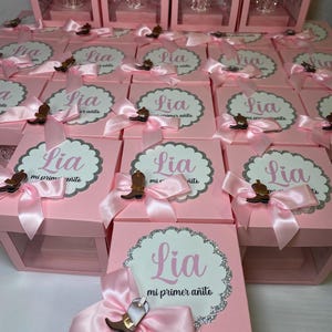 May include: Pink gift boxes with clear lids, each containing a small glass crown. The boxes are decorated with pink bows and a silver glitter design. The boxes have a label that says "Lia mi primer a&ntilde;ito" which translates to "Lia my first year".