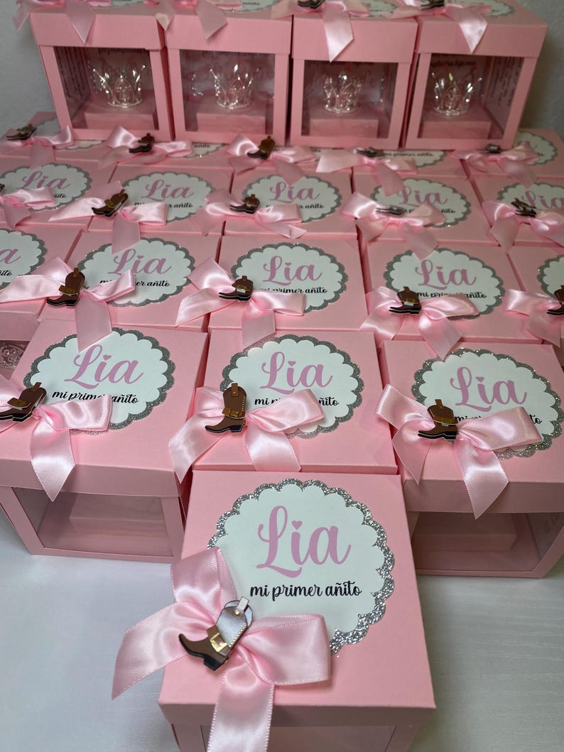 May include: Pink gift boxes with clear lids, each containing a small glass crown. The boxes are decorated with pink bows and a silver glitter design. The boxes have a label that says "Lia mi primer a&ntilde;ito" which translates to "Lia my first year".