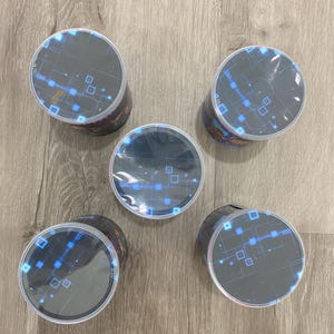 May include: Five cylindrical containers with black sides and clear, circular tops. The tops feature a blue and white digital design with squares and lines. The containers are arranged on a light wood-grain surface.