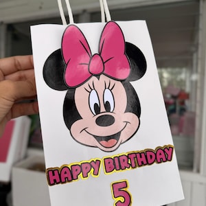 May include: White gift bag featuring a Minnie Mouse design with a pink bow. The bag has the text "HAPPY BIRTHDAY" in gold and pink, with the number "5" below. The bag has white handles.