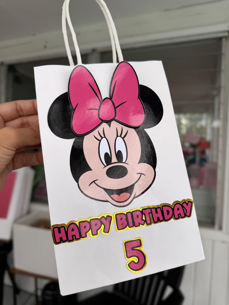 May include: White gift bag featuring a Minnie Mouse design with a pink bow. The bag has the text "HAPPY BIRTHDAY" in gold and pink, with the number "5" below. The bag has white handles.