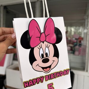 May include: White gift bag featuring a Minnie Mouse design with a pink bow and black ears. The bag has the text "HAPPY BIRTHDAY 5" in pink and gold. The bag has white rope handles.