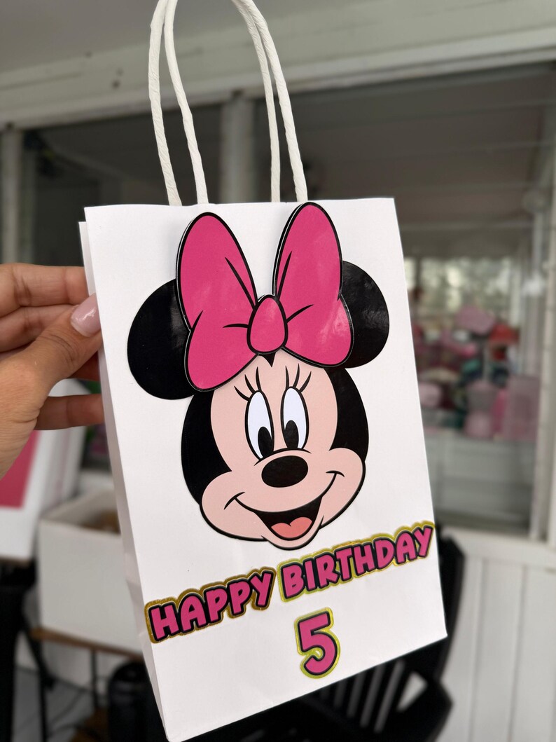 May include: White gift bag featuring a Minnie Mouse design with a pink bow and black ears. The bag has the text "HAPPY BIRTHDAY 5" in pink and gold. The bag has white rope handles.