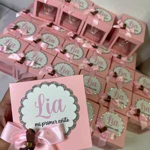 May include: Pink favor boxes with a clear window, each decorated with a pink bow and a silver glitter circle with the name "Lia" and the text "mi primer a&ntilde;ito".