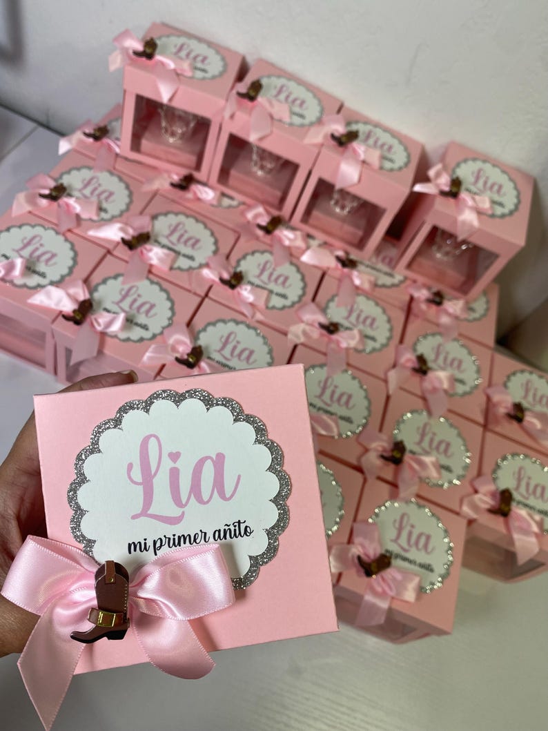 May include: Pink favor boxes with a clear window, each decorated with a pink bow and a silver glitter circle with the name "Lia" and the text "mi primer a&ntilde;ito".