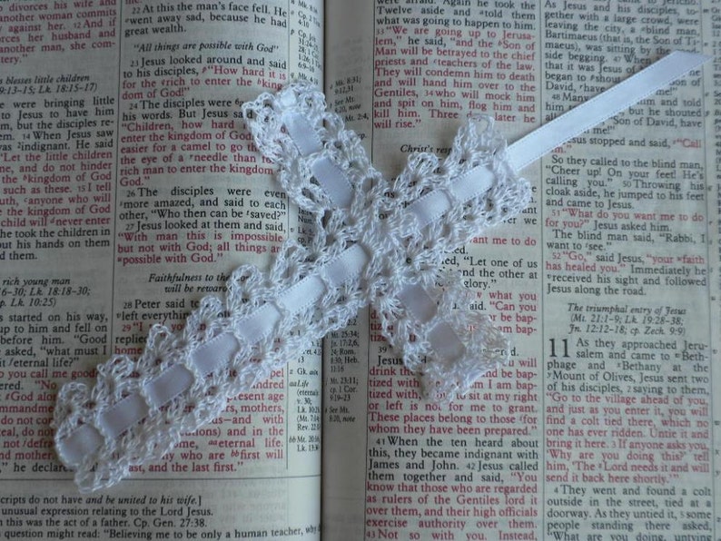 May include: A white crocheted lace bookmark with a white satin ribbon. The bookmark is placed on an open Bible with text about Jesus. The text reads, "All things are possible with God."