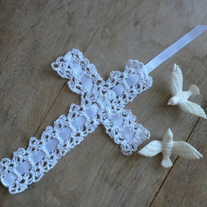 May include: White crocheted cross with a white satin ribbon attached. Two white ceramic doves are on a wooden surface.