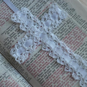 May include: A white crocheted bookmark with a white ribbon attached to a page in an open book. The book is open to a passage from the Bible.