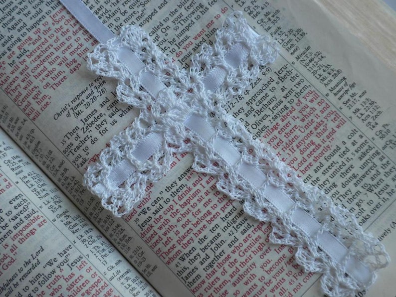 May include: A white crocheted bookmark with a white ribbon attached to a page in an open book. The book is open to a passage from the Bible.
