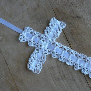 May include: White crocheted cross with a white satin ribbon attached. The cross is on a wooden surface.