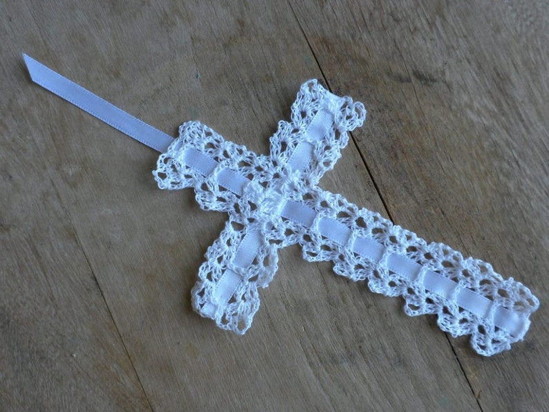 May include: White crocheted cross with a white satin ribbon attached. The cross is on a wooden surface.