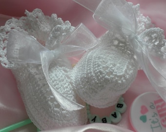 May include: Two white crocheted baby booties with white satin ribbon bows. The booties are on a pink satin surface with a white and pink "It's a Girl" sign.