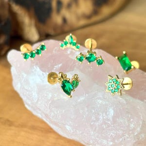 Może przedstawiać: Kolekcja złotych kolczyków sztyftów z różnymi zielonymi kamieniami szlachetnymi, w tym klastrami, sercami i kwadratem. Kolczyki są prezentowane na jasnoróżowym, chropowatym kamieniu, na tle rozmytego drewnianego tła.