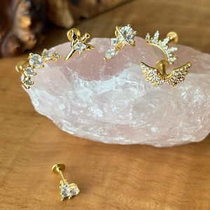 May include: Gold-toned earrings with clear, sparkling gemstones. The collection includes a star, a crescent, and other decorative designs. The earrings are displayed on a pink stone, showcasing their intricate details and elegant style.