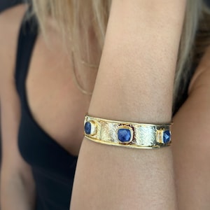 May include: A gold-toned cuff bracelet featuring a textured, hammered metal surface. The bracelet is accented with three square-cut, faceted blue gemstones set in individual gold-toned bezels spaced evenly along the center of the band.