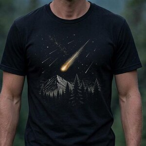 Peut inclure: Un t-shirt noir à manches courtes arborant une illustration d'une étoile filante jaune vif traversant un ciel nocturne étoilé au-dessus d'une chaîne de montagnes et d'une forêt dense de pins, avec des détails en lignes blanches et grises.