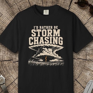 Puede incluir: Una camiseta negra de manga corta con el texto I'D RATHER BE STORM CHASING en color beige sobre una ilustración gráfica de una gran nube de tormenta con un rayo impactando en un campo de hierba.
