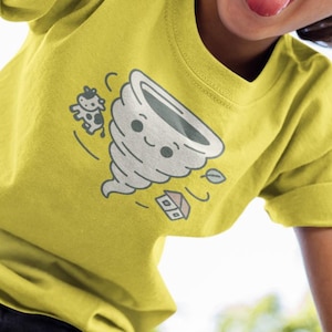 Puede incluir: Una camiseta amarilla con un diseño de tornado de dibujos animados, una vaca y una casa. La camiseta la lleva un niño con el pelo rizado castaño y vaqueros azules. El niño sonríe y saca la lengua.