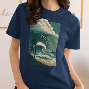 May include: Navy blue t-shirt featuring a coastal scene with a stormy sky, lightning, crashing waves, palm trees, and a lighthouse. The graphic is a rectangular design on the front of the shirt.